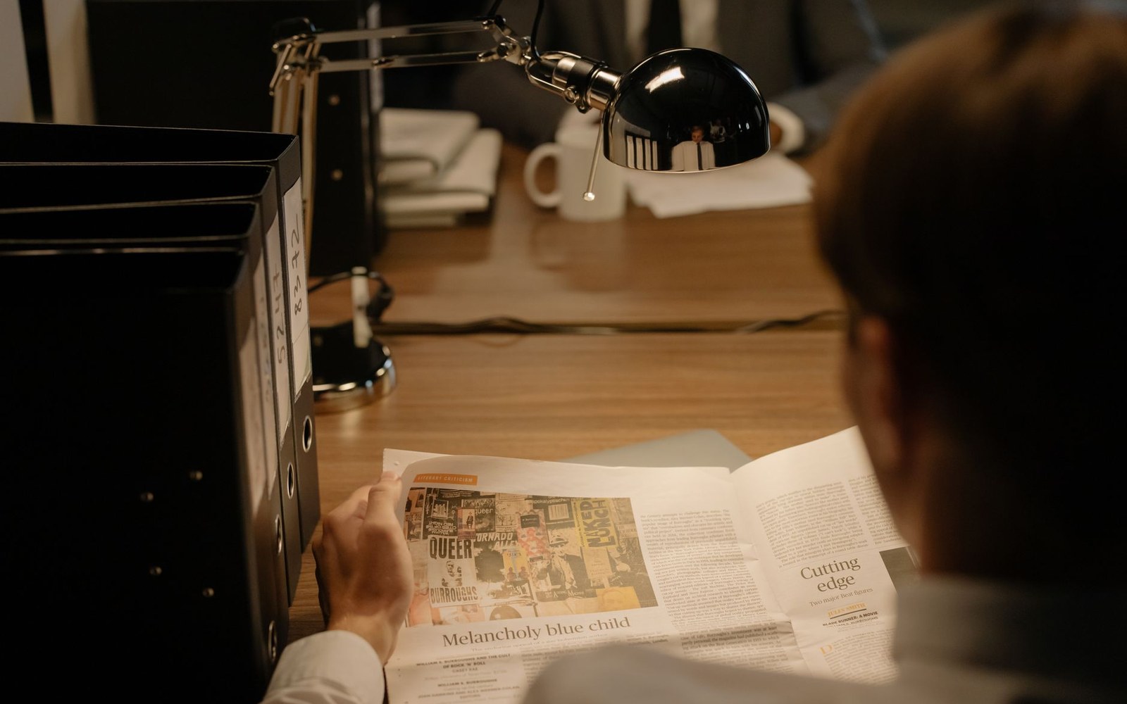 A person reads a newspaper article at a desk lamp-lit office desk with binders and a mug