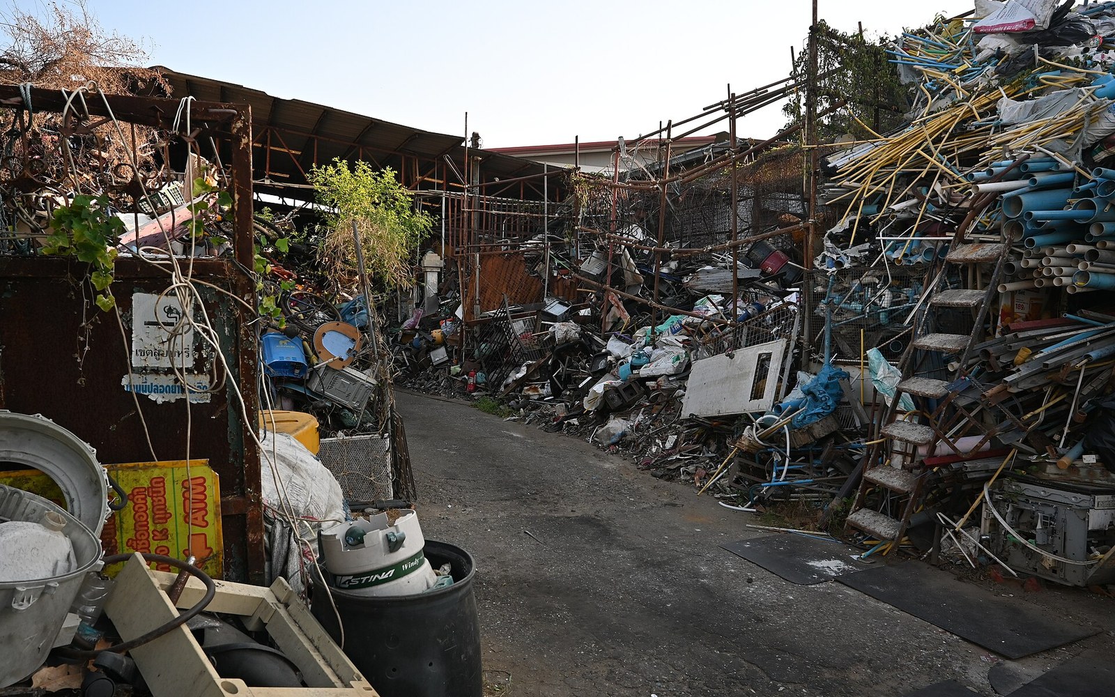 Piles of scrap metal and discarded pipes line a narrow junkyard pathway